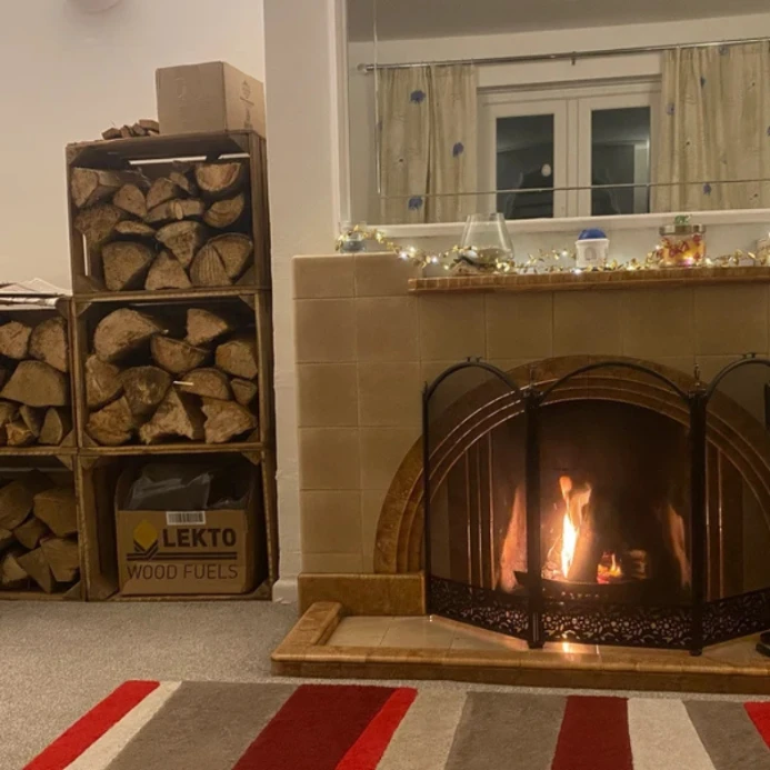 Three wooden crates stacked in a column and filled with split logs beside a lit open fire with a fireguard